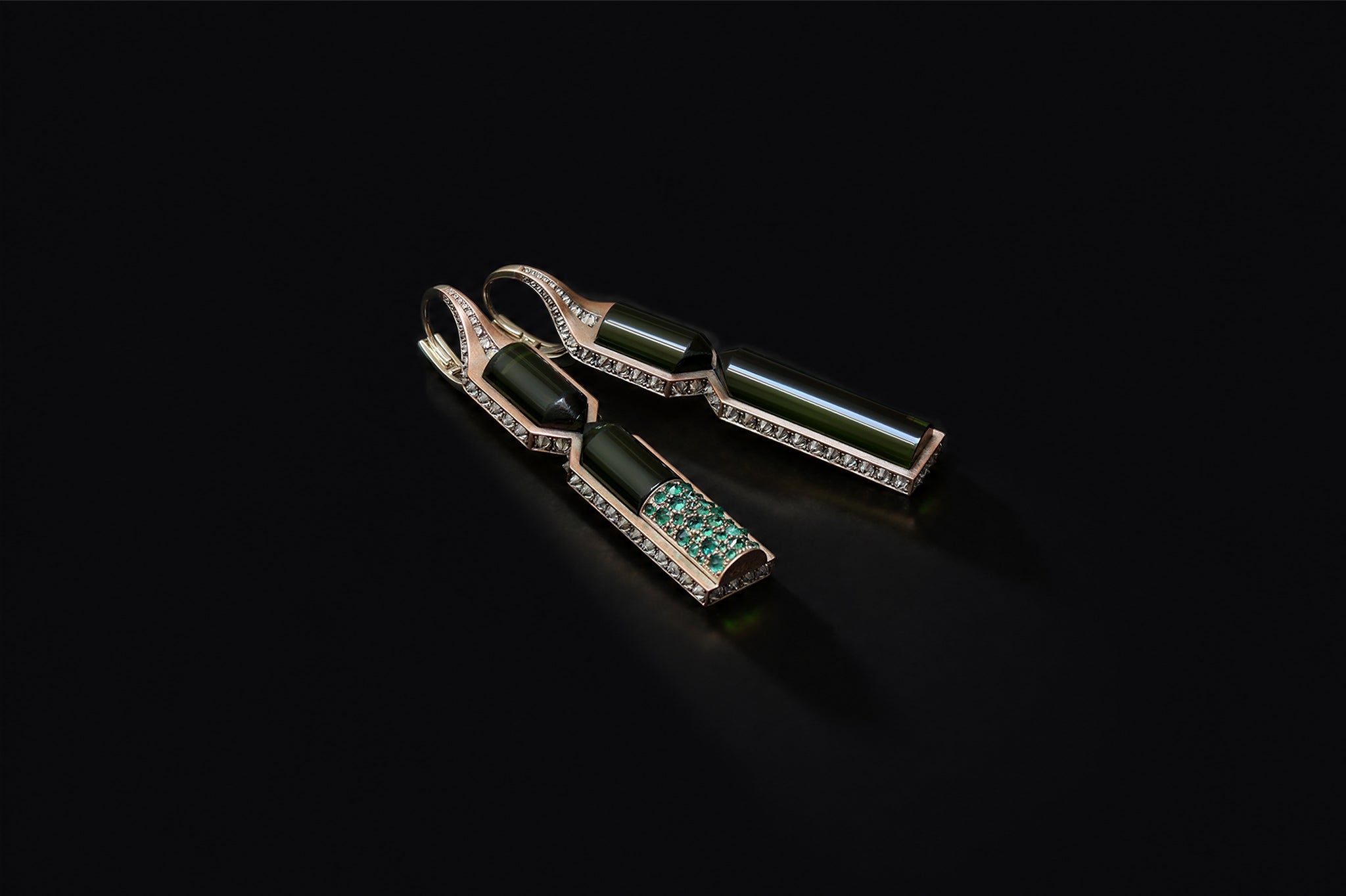 Aleutian Precipice Earrings on a black background, the elongated dark green tourmaline batons edged with diamonds, one revealing a pave of emeralds along its length.